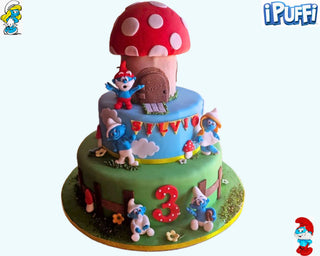 Torta a tema Villaggio dei Puffi a due piani, decorata con Grande Puffo, Puffetta e altri personaggi in pasta di zucchero, con casetta a fungo e dettagli colorati.